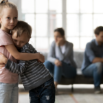 Little girl hugs brother as parents avoid each other in the background
